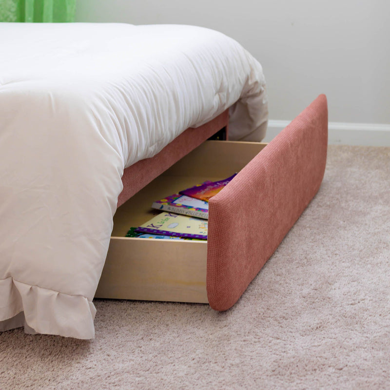 Under-bed storage drawer in a pink upholstered bed frame filled with children's books, white duvet and beige carpeted bedroom floor