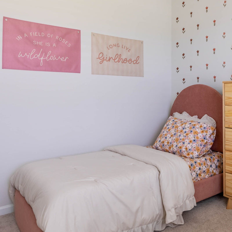 Girls bedroom with neutral beige duvet, floral pillowcase and blush pink upholstered headboard, pink and cream wall banners reading "wildflower" and "girlhood" and scattered flower wall decals — feminine bedroom decor inspiration