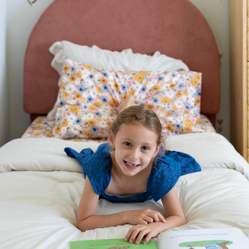 Girl in blue dress lying on bed with floral pillows, reading a book and smiling.
