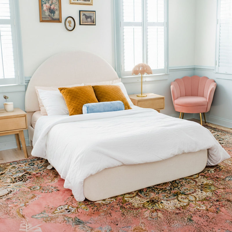 Pastel bedroom with curved upholstered bed, white linens, mustard velvet pillows, blue bolster, pink shell chair, wooden nightstand, gold lamp, shuttered windows and patterned pink boho rug