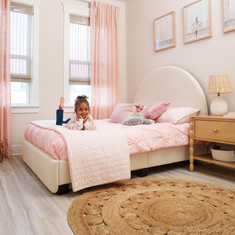 Pink girl's bedroom with tufted pink bedding, round upholstered headboard, blush curtains, jute rug, wicker lamp and smiling child on bed, modern kids' room decor