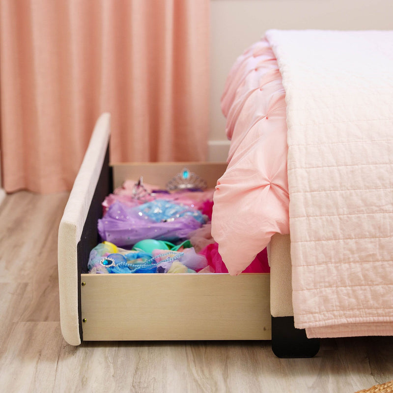 Under-bed storage drawer filled with colorful dress-up costumes beneath a tufted pink bed and soft pink quilt in a girl's bedroom with matching pink curtains