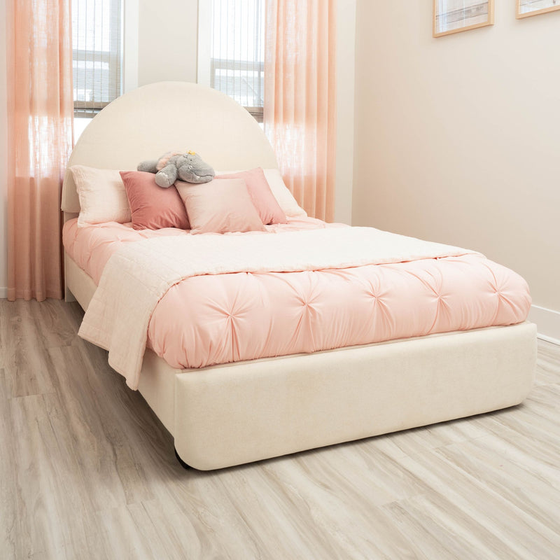 Pastel pink bedroom with soft pink bedding, rounded upholstered headboard, peach sheer curtains, stuffed hippo toy and light wood flooring