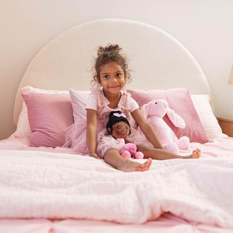 Smiling young girl sitting on a pink bed with a plush bunny and doll, surrounded by soft pillows and pastel children's bedroom decor