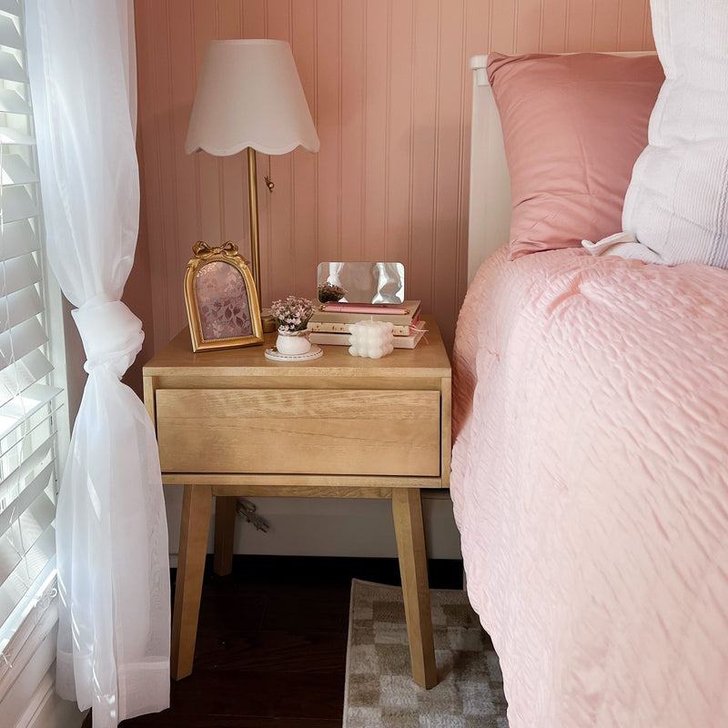 Pink bedroom nightstand with scalloped white table lamp, wooden bedside table, pink quilted bedding, sheer white curtains and feminine decor