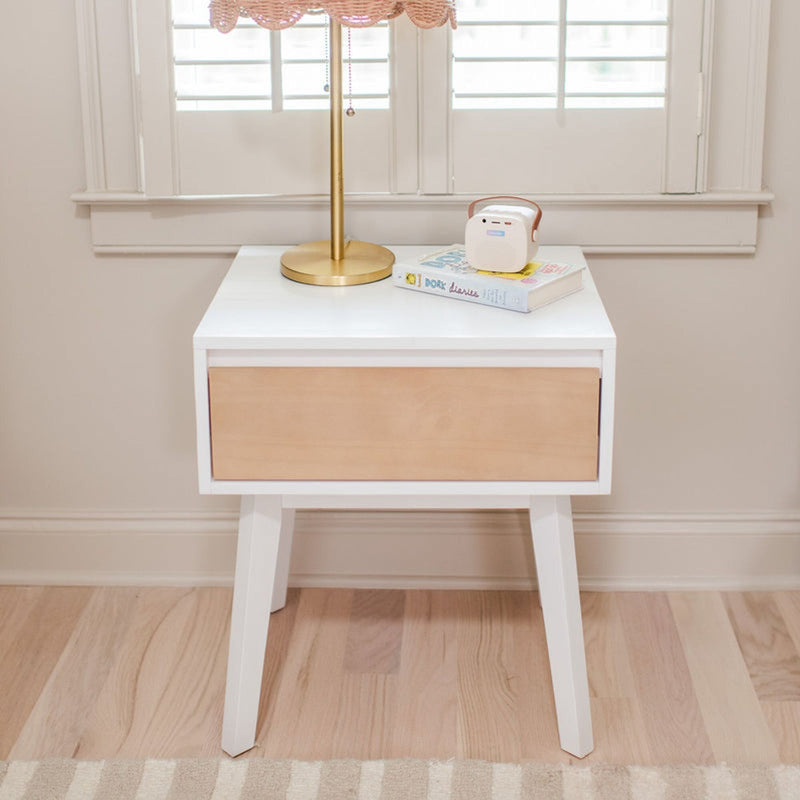 Modern mid-century white and natural wood nightstand with angled legs, brass lamp with pink shade, children's book and small white portable speaker on hardwood floor by shuttered window, nursery bedside decor