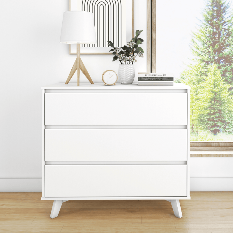 Minimalist white three-drawer dresser in a Scandinavian bedroom with a wooden tripod table lamp, small clock, ceramic vase of greenery, stacked books, hardwood floor and sunlit window showing pine trees.