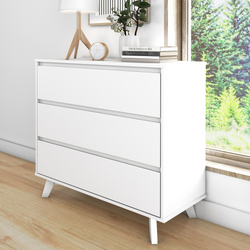 Minimalist white 3-drawer dresser / chest of drawers with angled wooden legs, styled with table lamp, round clock, potted plant and books on oak floor by sunny window overlooking green trees