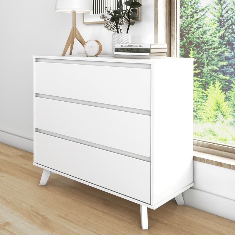 White modern three-drawer dresser with angled wooden legs in a minimalist bedroom, styled with a table lamp, clock, stacked books and plant by a sunny window