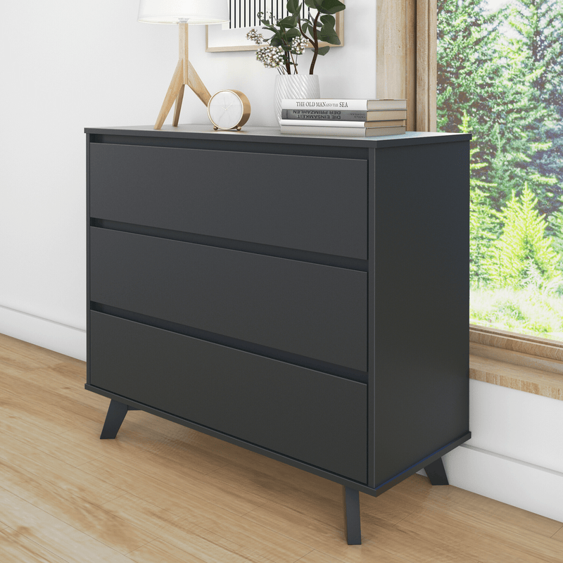 Modern matte-black three-drawer dresser with angled mid-century legs, styled with lamp, clock, books and potted plant beside a window overlooking green trees