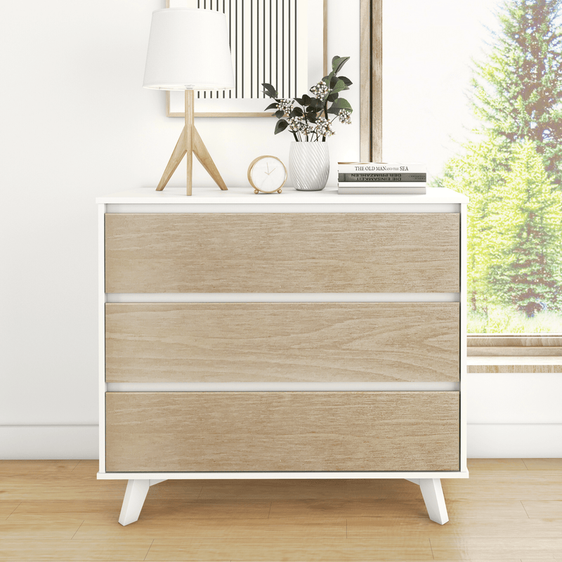 Modern Scandinavian three-drawer dresser with light oak fronts and white frame styled with a table lamp, clock, potted plant and books by a sunlit window