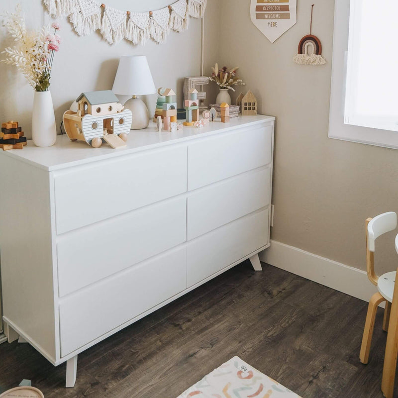 White modern dresser in neutral boho nursery with wooden toys, lamp, vases and rainbow wall hanging, kids room storage