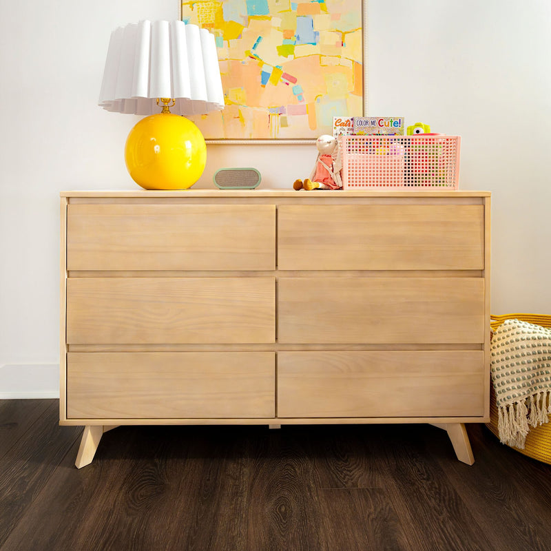 Light wood mid-century dresser with bright yellow globe table lamp, pastel abstract wall art, pink toy storage basket and knit throw on dark hardwood floor