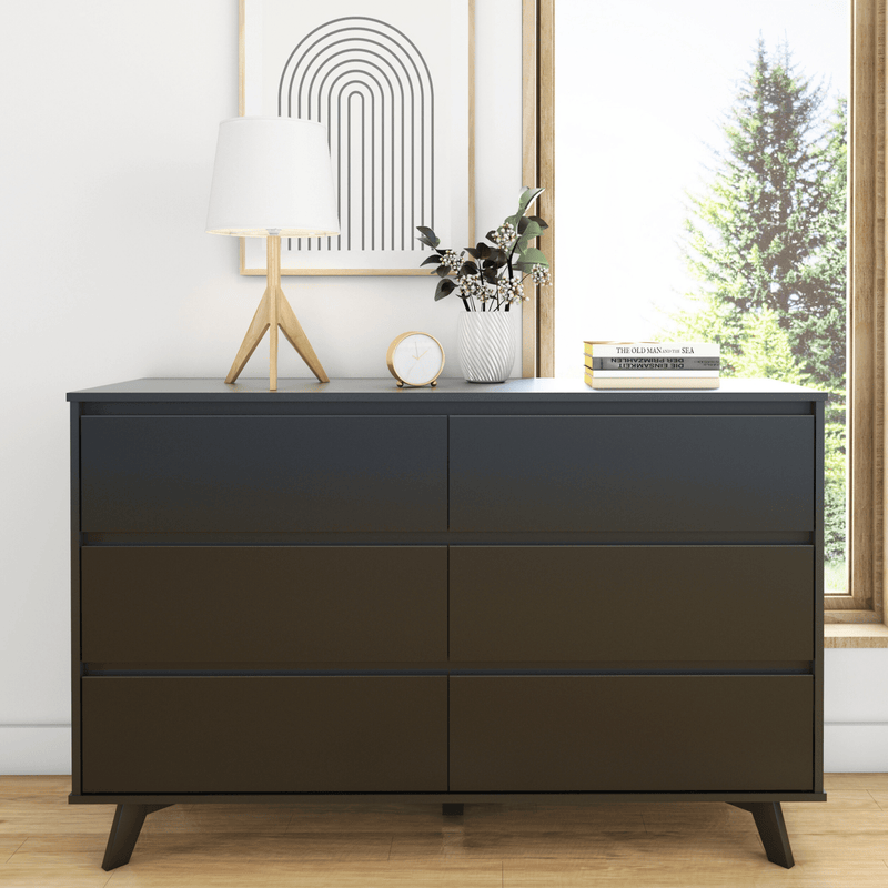 Modern black dresser styled with a wooden tripod lamp and white shade, gold alarm clock, white vase of greenery, minimalist arch print and stacked books beside a window with pine tree view