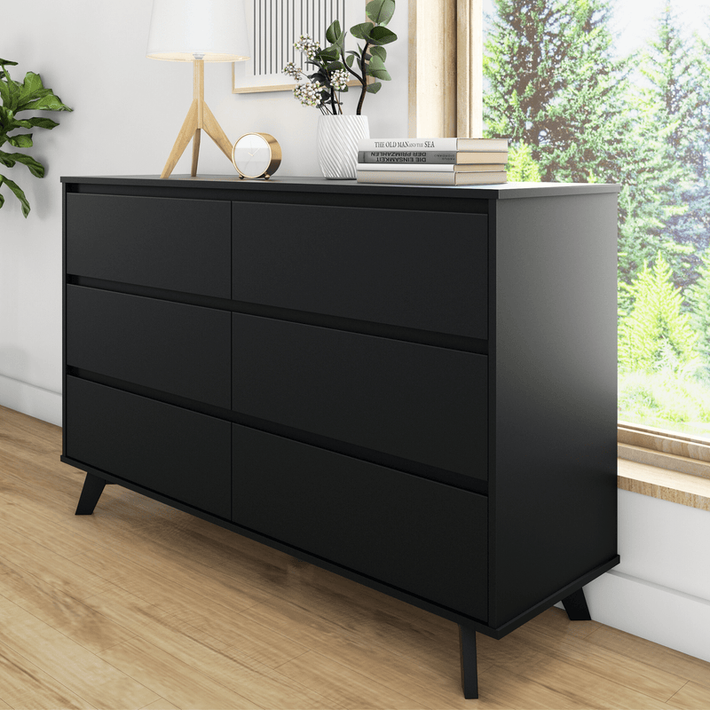 Modern black dresser in a minimalist Scandinavian bedroom with wooden tripod lamp, gold clock, white vase with greenery and stacked books by a sunlit window
