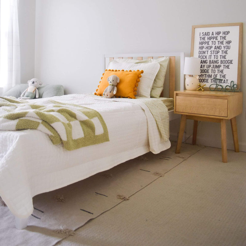 Cozy neutral children's bedroom nursery with white bed, green check bedding, orange pom-pom pillow, plush toys, wooden nightstand, lamp and framed print