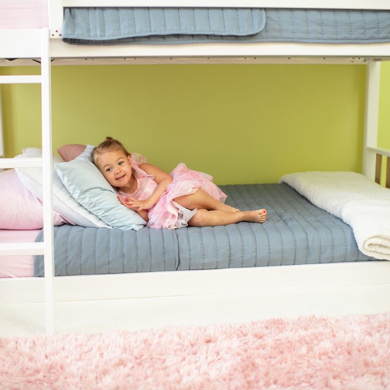 Young girl in pink dress lying on lower bunk bed with blue quilt and pastel pillows in a cozy kids' bedroom with pink rug