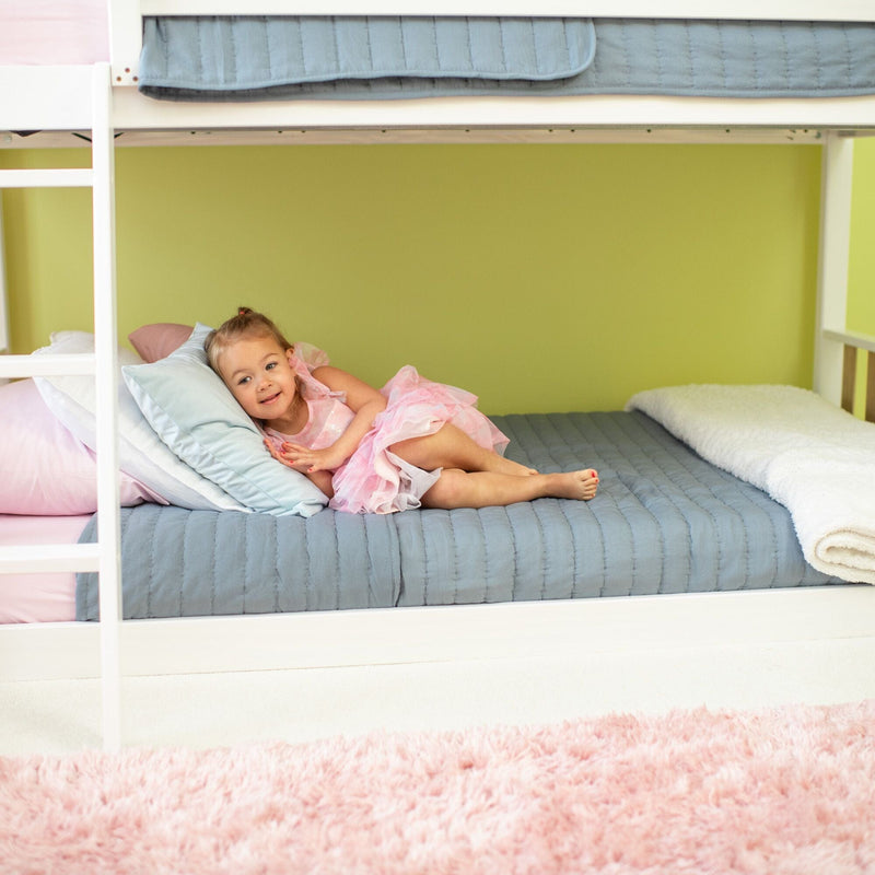 Young girl in a pink dress lying on the lower bunk bed with gray quilt and pastel pillows in a bright kids' bedroom with a pink shag rug