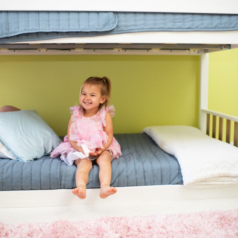 Smiling toddler girl in a pink dress sitting on a blue-quilted bottom bunk bed in a bright pastel kids' bedroom with a white blanket and pink rug