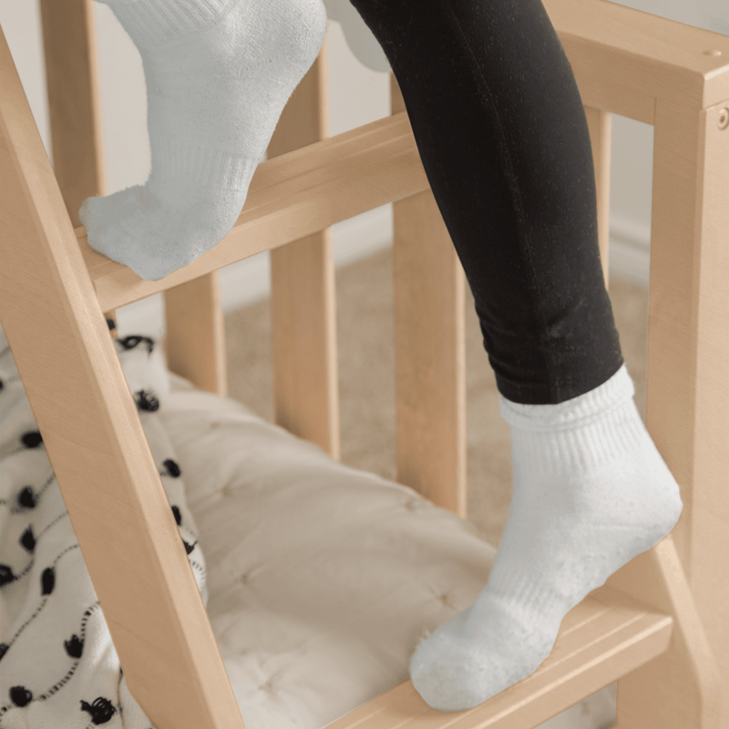 Close-up of toddler's feet in white socks and black leggings resting on a natural wood crib rail with soft nursery bedding