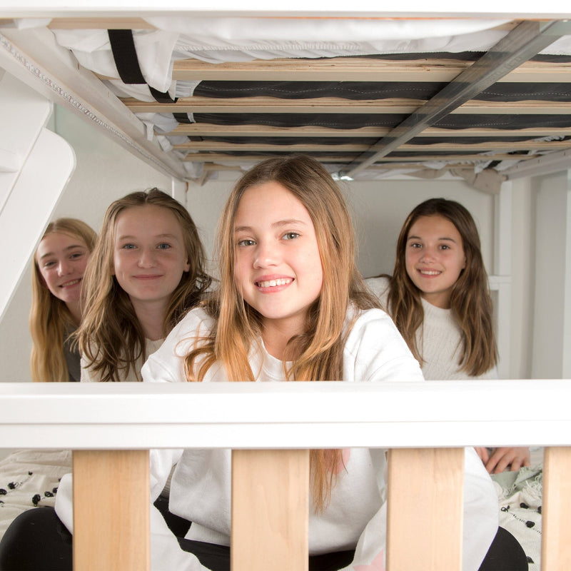 Group of four smiling girls at a sleepover sitting on a lower bunk bed in a bright cozy bedroom with white bedding and a wooden frame