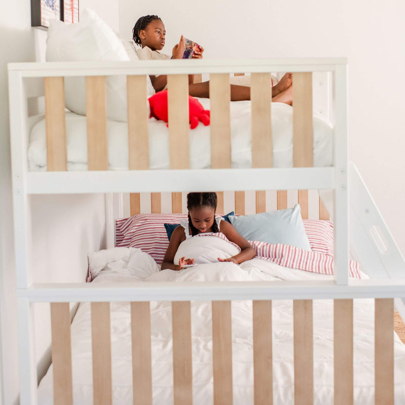 Two children on a white wooden bunk bed in a cozy kids' bedroom — top child reading a book, bottom child using a tablet