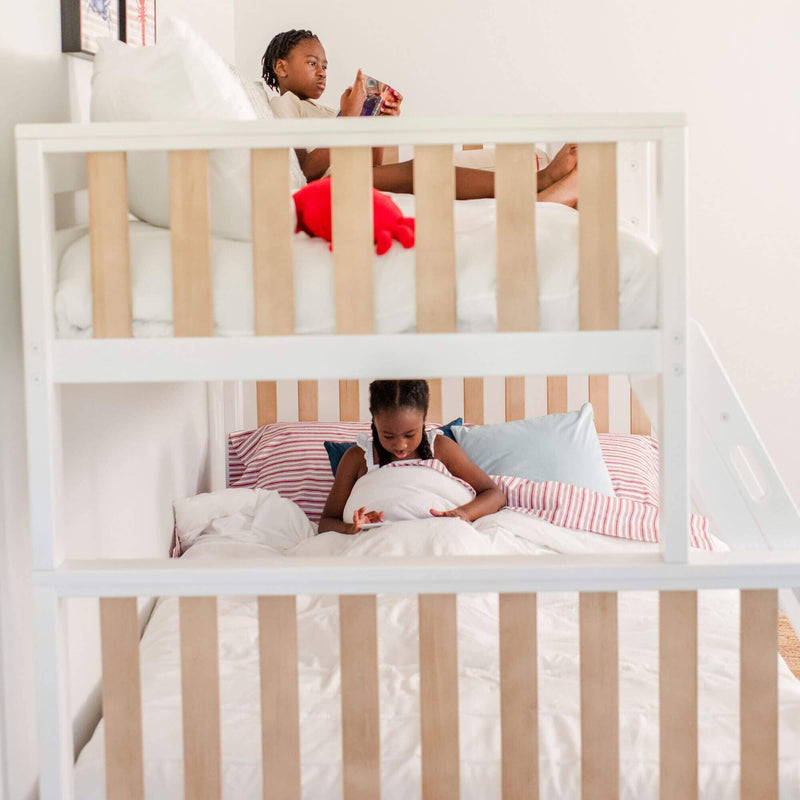 Two children on a white wooden bunk bed in a cozy kids' bedroom — top child reading a book, bottom child using a tablet