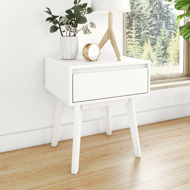 White modern bedside table nightstand with single drawer and wooden legs, lamp, vase with greenery and gold alarm clock on hardwood floor by window with forest view