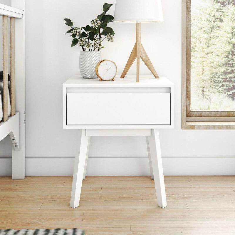 White Scandinavian nightstand with a drawer, decorative plants, modern lamp, and clock in a bright bedroom setting.