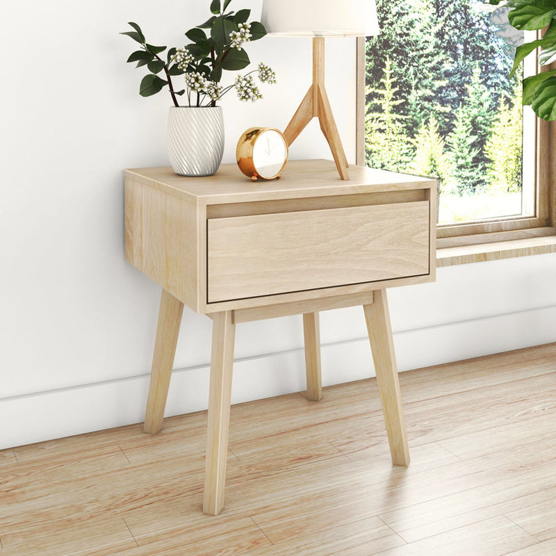 Scandinavian wooden nightstand with white vase and green branches, gold alarm clock and tripod lamp by window with forest view