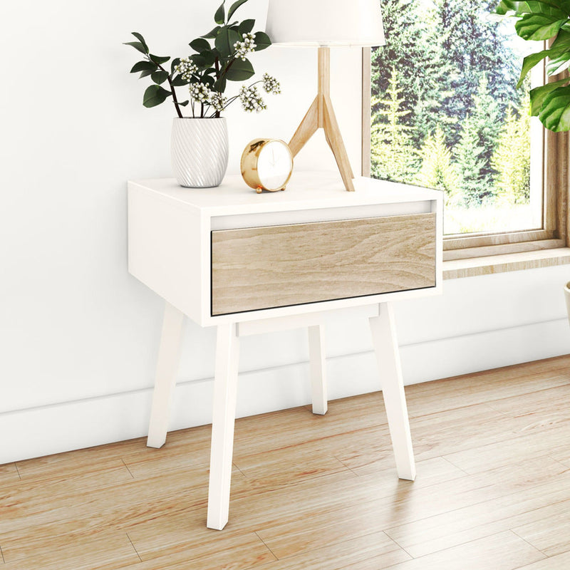 Minimalist Scandinavian bedroom with white floating bedside table and wooden drawer, tripod table lamp, white ceramic vase with greenery, gold alarm clock, hardwood floor and sunlit forest view through window