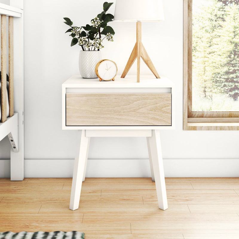 Minimal Scandinavian bedside table with wooden tripod lamp, white ceramic vase of greenery and gold alarm clock by sunlit window