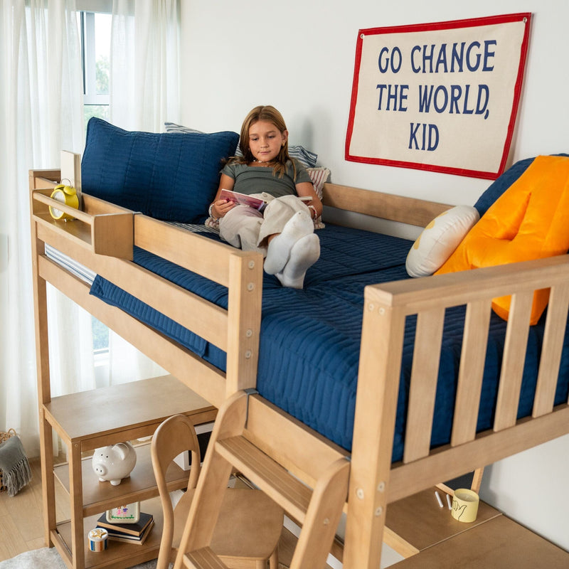 Child reading on a wooden loft bunk bed with navy bedding, blue pillows, yellow alarm clock and motivational "Go Change the World, Kid" wall banner in a bright kids' bedroom