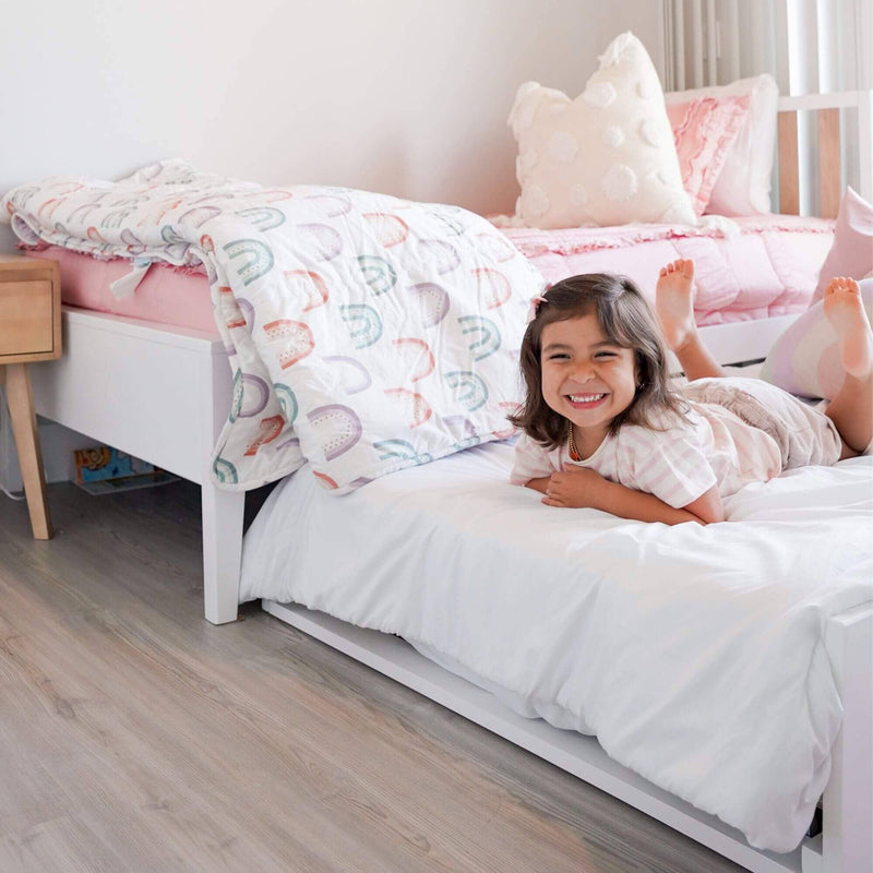 Smiling child on a white bed with pastel rainbow quilt and pink children's bedding in a modern kids bedroom with decorative pillows and wood-look flooring