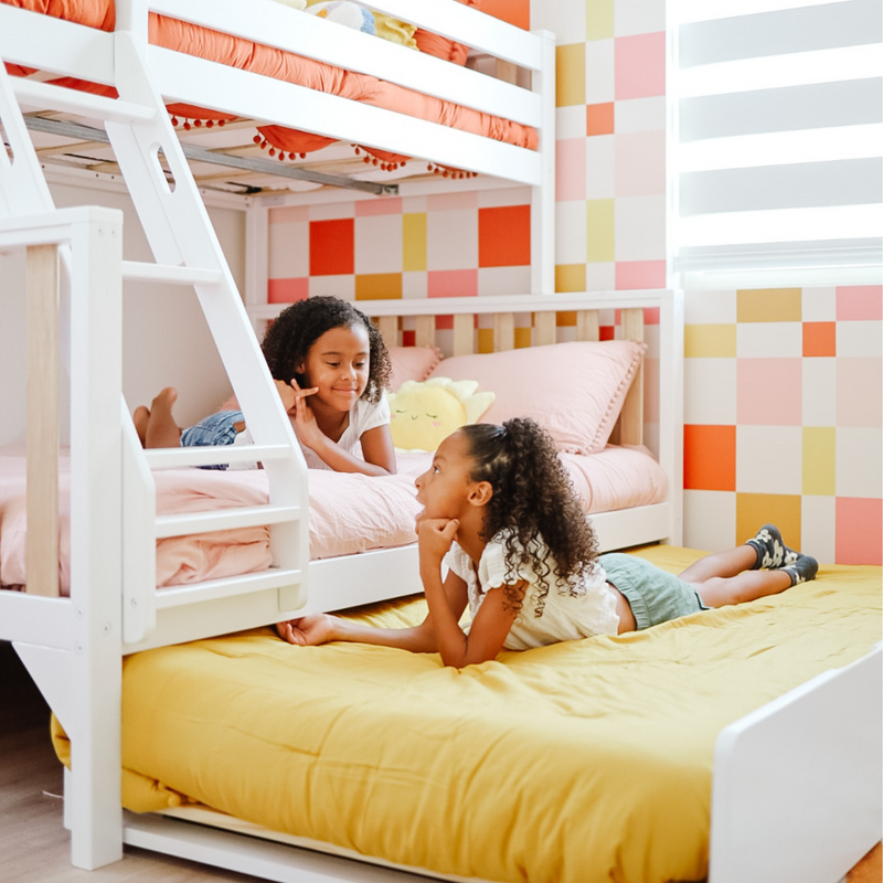 Two children playing on a lower bunk in a bright kids bedroom with a white bunk bed, pink bedding, yellow pull-out mattress and colorful checkered wallpaper.