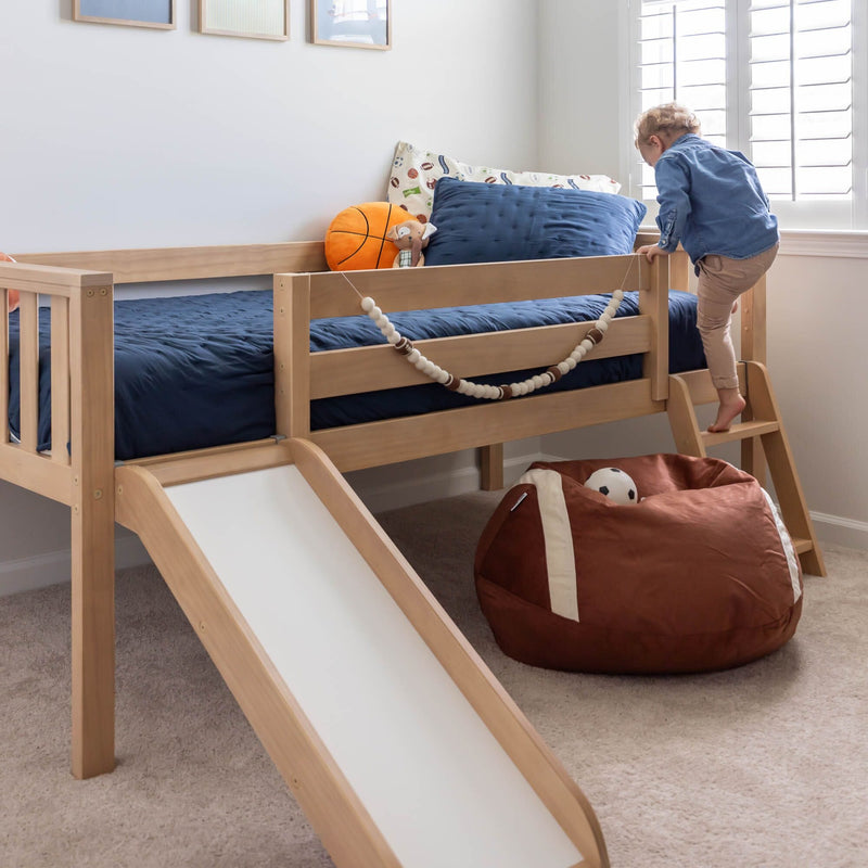Wooden kids loft bed with slide and ladder, navy bedding and sports-themed pillows with toddler climbing in bright bedroom