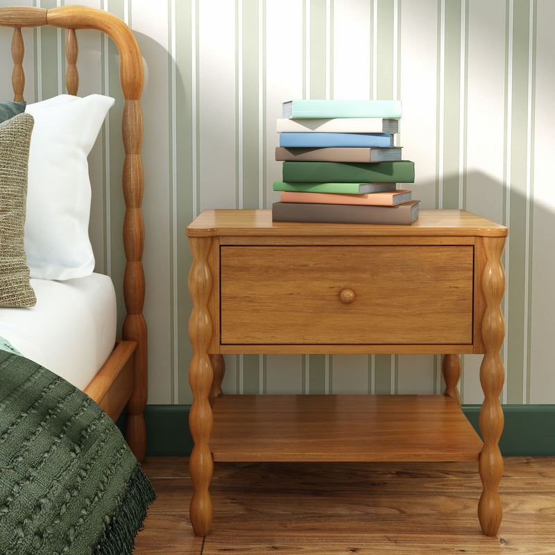Wooden bedside table nightstand with drawer and stack of books beside spindle wood bed frame, striped wallpaper and green throw blanket in cozy bedroom