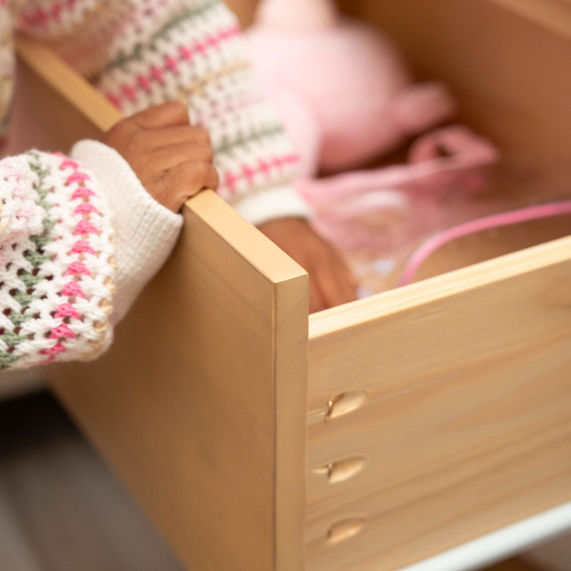 Toddler's hand in a pastel knitted sweater opening a wooden dresser drawer filled with pink toys — nursery storage and organization