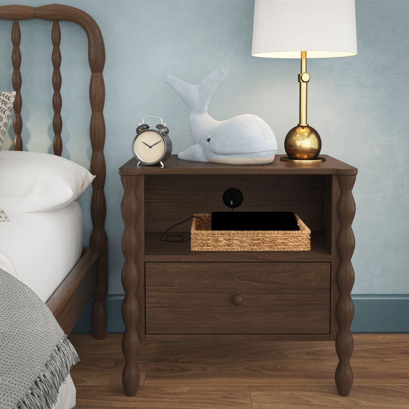 Dark wood nightstand with brass table lamp, alarm clock, blue whale plush toy and wicker tray holding a charging tablet against a light blue wall