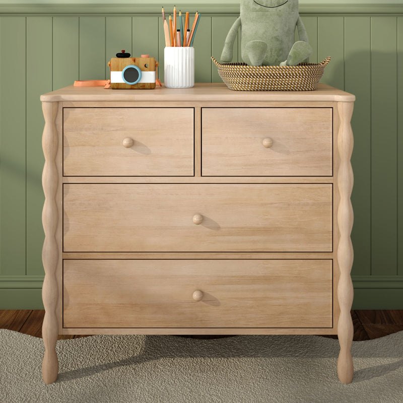 Light wood nursery dresser against green paneled wall with woven basket holding plush frog, white pencil vase and wooden toy camera — children's room storage and decor