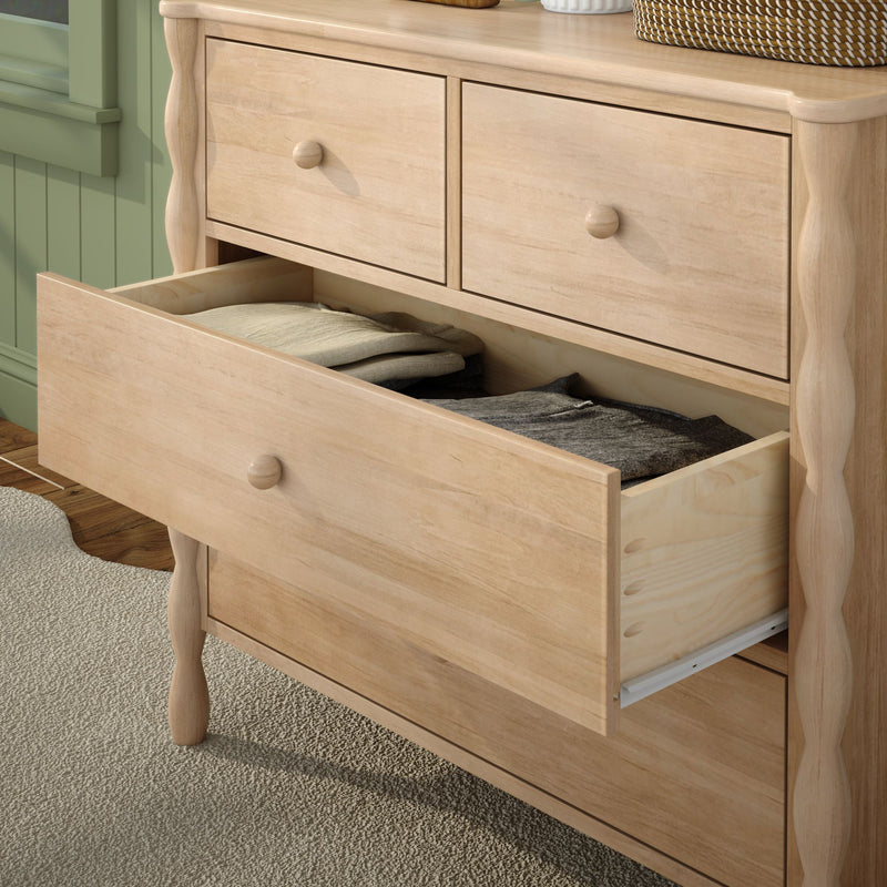 Light natural wood dresser with turned legs and an open drawer revealing folded clothes, bedroom storage chest