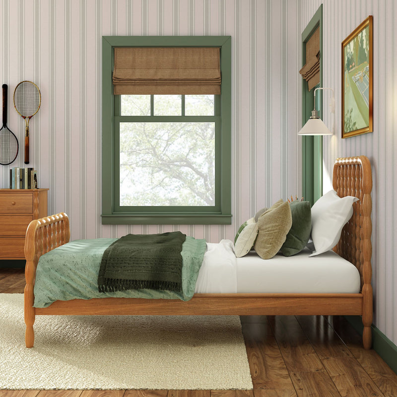 Cozy rustic bedroom interior with wooden bed frame, green bedding and pillows, green-trimmed window with brown Roman shade, striped wallpaper, wooden dresser and wall art, natural light