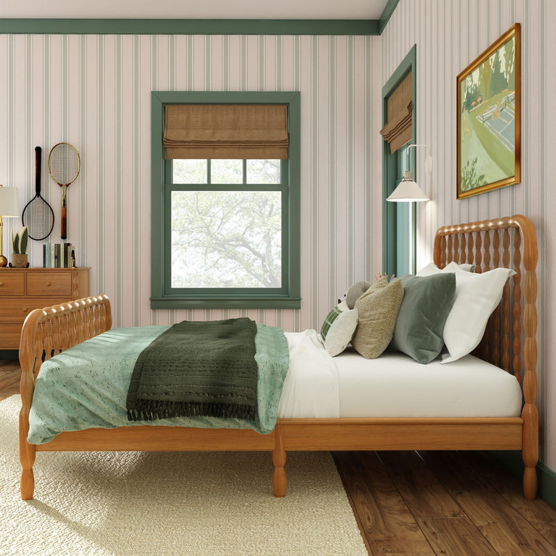 Cozy bedroom interior with wooden carved bed frame, green bedding and pillows, dark green throw, striped wallpaper, green-trimmed window with brown Roman shade, wooden dresser with books and vintage tennis rackets, wall lamp and framed artwork