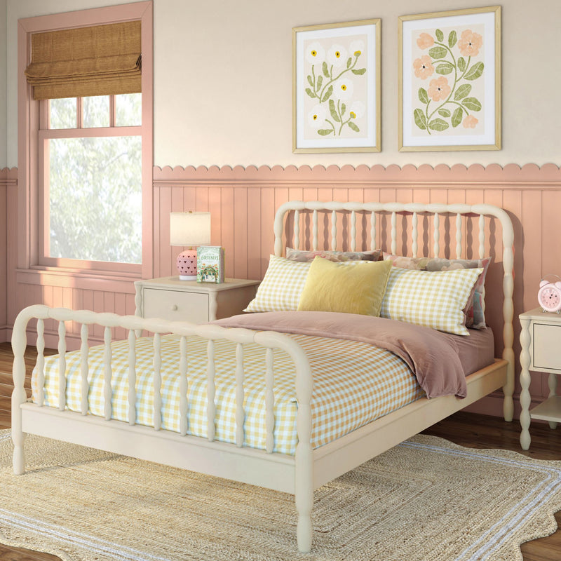Pastel pink bedroom with cream spindle bed, yellow gingham bedding, scalloped wainscoting, floral framed prints, roman shade and bedside lamp