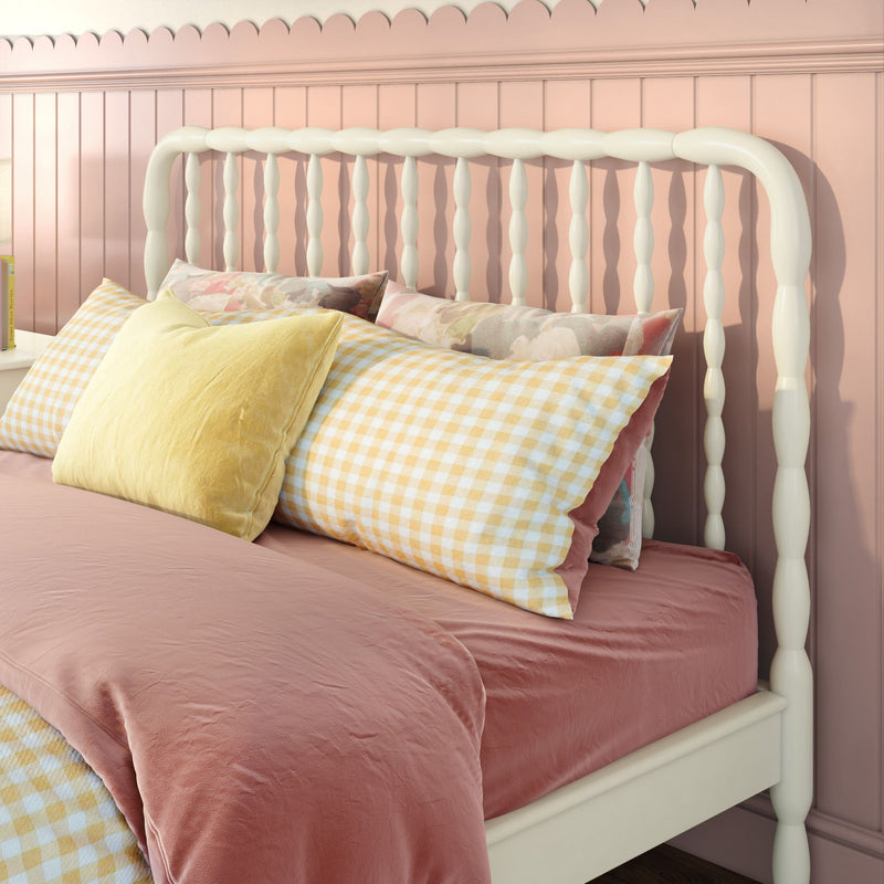 White spindle headboard on pink scalloped wall with pastel gingham pillows and blush velvet bedding — cozy nursery bedroom decor