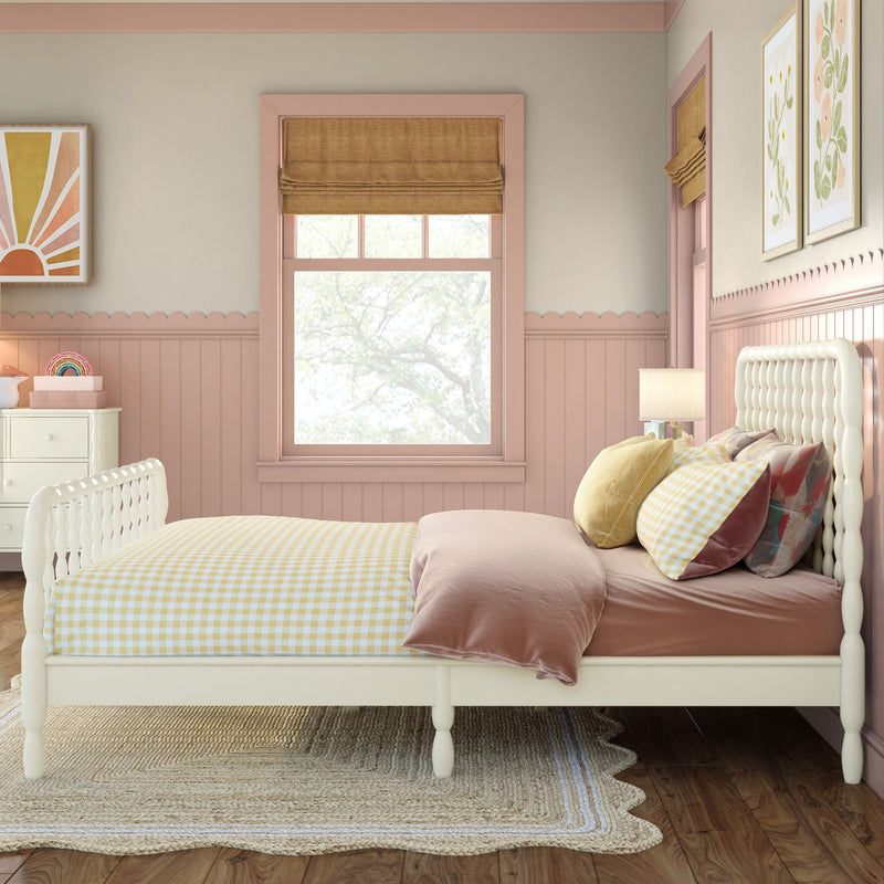 Cozy pastel pink girls' bedroom with white spindle bed, yellow gingham bedding, layered decorative pillows, scalloped wainscoting, roman shade and framed botanical art