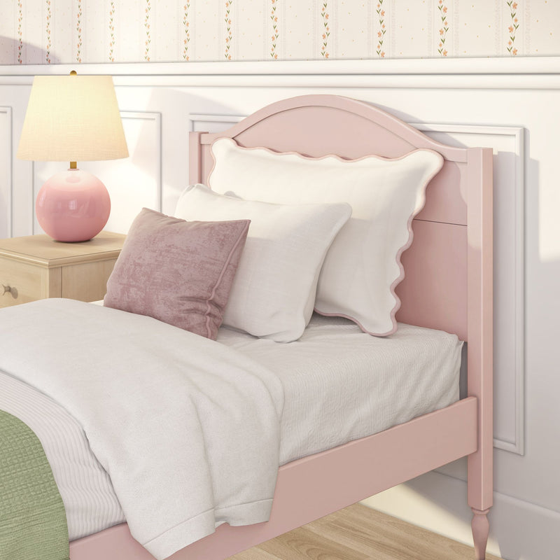 Pastel girls bedroom close-up with pink scalloped headboard, white bedding, velvet pink throw pillows, pink ceramic bedside lamp on wooden nightstand and floral wallpaper accent