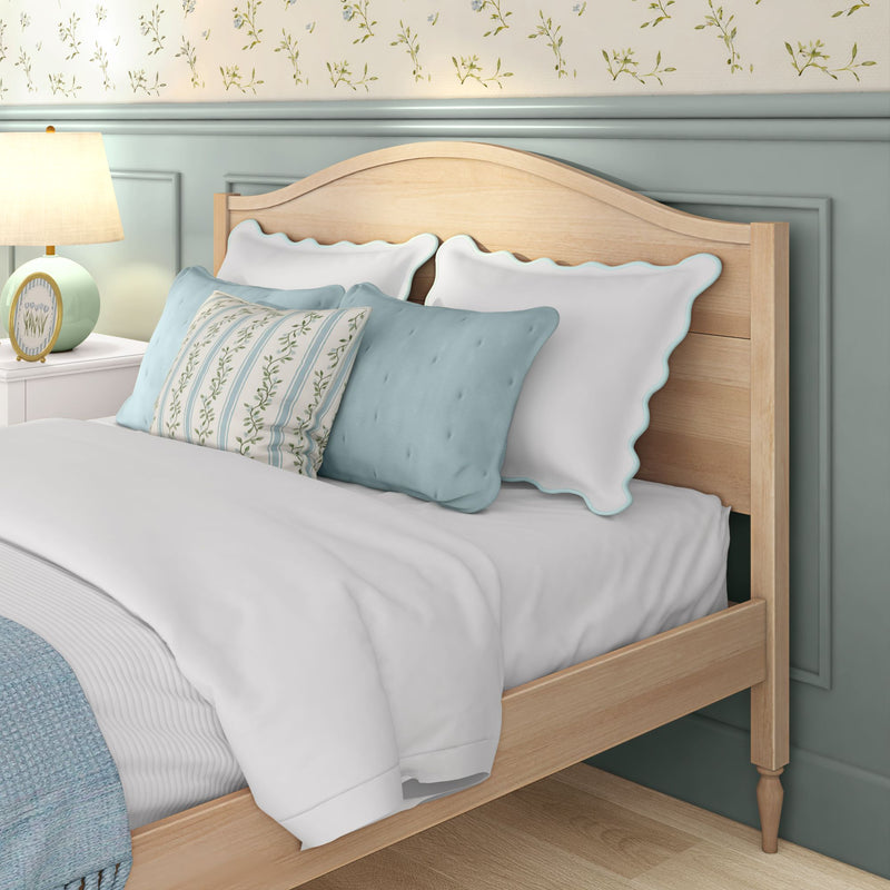 Cozy coastal bedroom close-up of natural wood bed frame with white scalloped pillows blue throw pillows floral wallpaper pastel wainscoting and bedside lamp