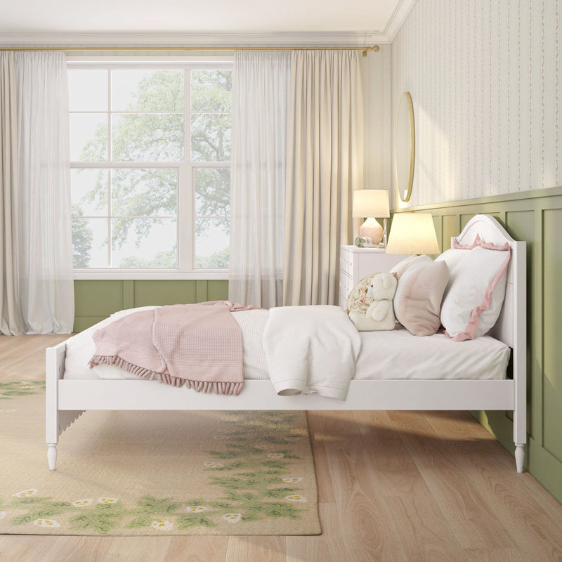 Pastel pink children's bedroom with white bed frame, pink quilt and pillows, plush teddy bear, bedside lamp, oval wall mirror, green wainscoting and large window with sheer curtains