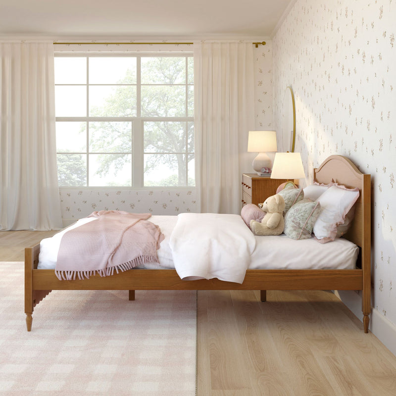Cozy pastel girl's bedroom with wooden bed frame, pink bedding and throw, teddy bear on bed, floral wallpaper, bedside lamp and large window with sheer white curtains